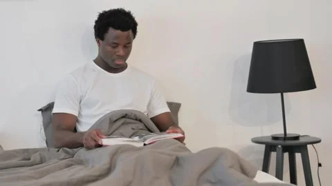 African Man Reading Book while Sitting in Bed Stockbeeldmateriaal 299756377