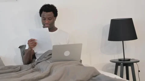 African Man Reading Documents while Working on Laptop while Sitting in Bed Stockbeeldmateriaal 299755337