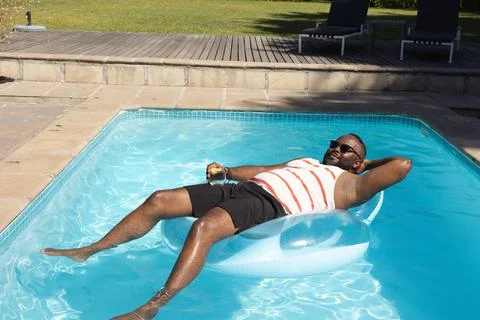 African man reclining on translucent ring in backyard pool holding clear glass, Stock Photos