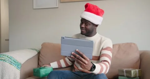 African man in Santa hat looking at tablet, lifting green gift showing it on Vidéo 343889102
