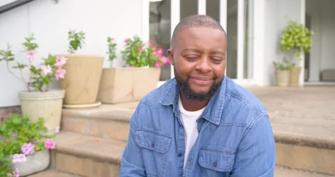 African man sitting on front steps, reacting to camera shift, smiling, laughing, Stock Footage 338326287