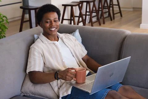 African man sitting on gray sofa at home holding peach mug and using silver Stock Photos