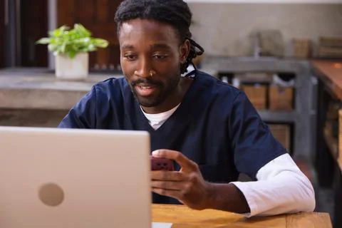 African man sitting at kitchen table wearing navy scrubs using silver laptop and Stock Photos
