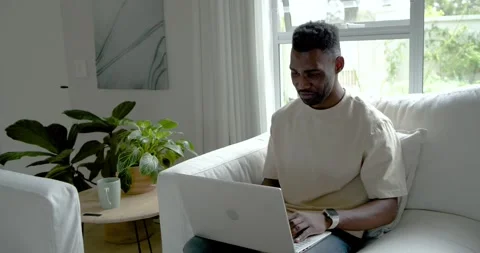African man sitting on light sofa in living room reading laptop alert, typing Stock Footage 339320963