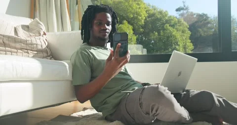 African man sitting on rug by sofa in green tee, typing silver laptop, answering Stock Footage 339163531