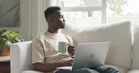 African man sitting on sofa, holding mug, typing on laptop, resting eyes while Stock Footage 339651767