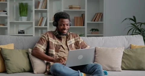 African man sitting on sofa at home, playing online game on laptop, winning. Stock Footage 253825531