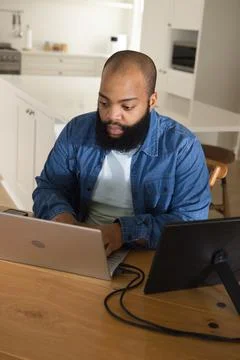 African man sitting, working on silver laptop, using tablet on stand with cables Fotos Stock