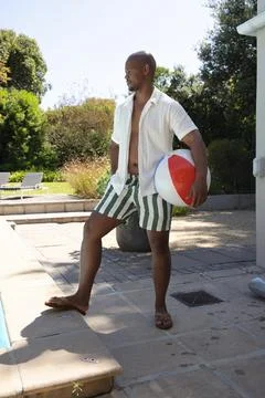 African man standing at backyard pool wearing open white shirt, striped trunks, Stock Photos