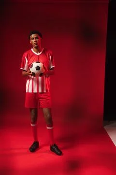 African man standing on red studio backdrop holding black-and-white soccer ball Stock Photos