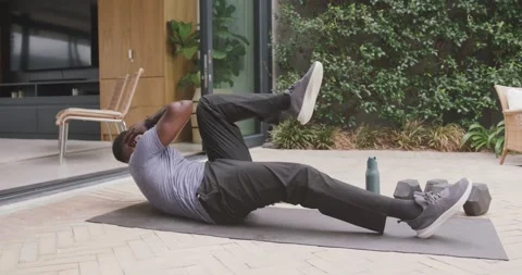 African man starting on back doing bicycle reps on patio mat with dumbbells, Stock Footage 340070485