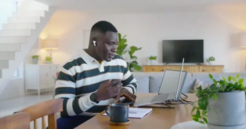 African man taking work call on phone, with earbuds, using laptop at home, Stock Footage 348224016