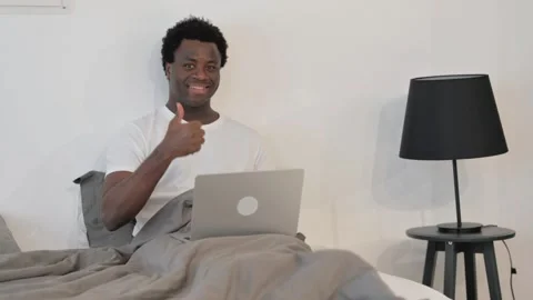 African Man with Thumbs Up Looking at Camera while in Bed with Laptop Video stock 299755638