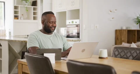 African man typing on laptop checking tablet smiling while reading message at Stock Footage 339321196