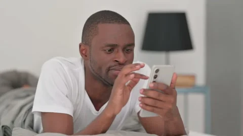 African Man Using Phone while Lying on Stomach in Bed Stock Footage 287901314