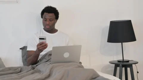 African Man Using Smartphone and Laptop while Sitting in Bed Vidéo 299755393
