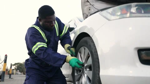 African mechanic in blue overalls changing a car tire in his workshop Stock Footage 346643232