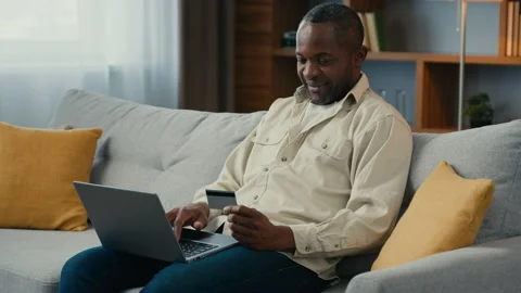 African middle aged man sitting on couch happy mature male customer paying for Stock Footage 229801040