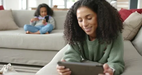 African mother tapping tablet while camera shifting focus, daughter on sofa Stock Footage 345098583
