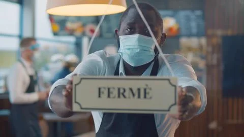 African waiter in safety mask turning ferme sign on french cafe glass door Stock Photos
