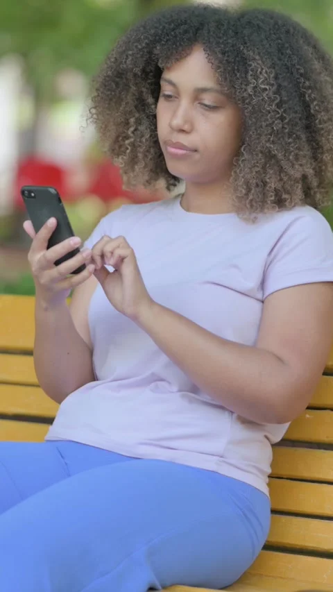 African Woman using Smartphone while Sitting Outdoor on a Bench, vertical Видео 326262838