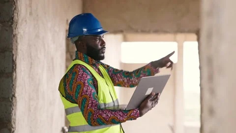 African worker in blue hard hat, vest reviews plans on laptop at site Video stock 346257686