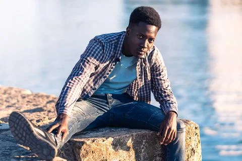 African young man posing facing the camera while sitting next to a river Stock Photos