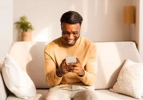 Afro Guy Using Cellphone Playing Games Sitting On Couch Indoor Stock Photos