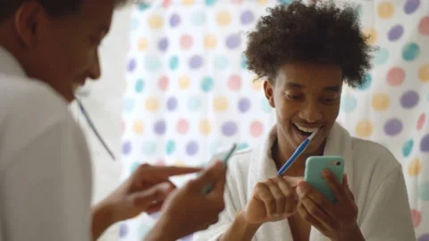 Afro man in bathrobe brushing teeth and using mobile phone in bathroom Stock Footage 141469232