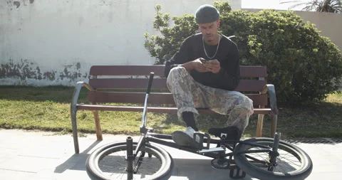Afro man sitting on a bench while looking at the mobile with his trial bike n Stock Footage 151484390