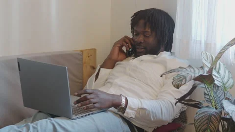 Afro man talks on the phone sitting on a sofa while working at home Stock Footage 147547887