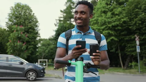 Afro man use smartphone stand with electric scooter and sipping hot drink to go Stock Footage 155682941