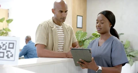 After man asking, African American nurse scrub-clad showing tablet to man at Stock Footage 336609407