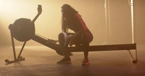 After rowing interval African American woman sitting on erg in gym holding water Stock Footage 339958253