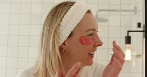 After selecting pink under-eye patches, mid adult woman applying them at vanity Stock Footage 337932828