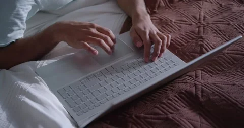 After settling on bed, hands are typing on silver laptop and using trackpad to Stock Footage 348442568