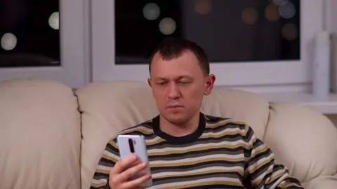 Against the background of a window with lights, a man sits with a phone in hands Stock Footage 132358644