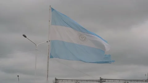 Aged Argentine flag waving in front of an industry with silos on a cloudy day Stock Footage 104925512
