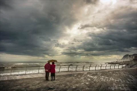 Aged couple staring at the horizon protected by umbrella under rainy sky Stock Photos