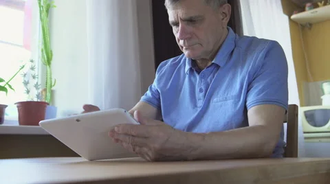 Aged man sits and uses a white tablet PC at home Видео 65504653