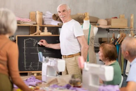 Aged teacher explaining pattern structure by chalkboard during sewing course for Stock Photos