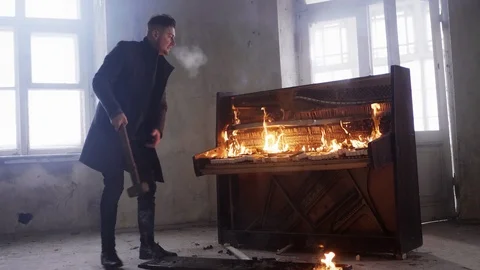 Aggressive man in grey coat with hammer in hand hits flaming retro wooden piano Stock Footage 95033838