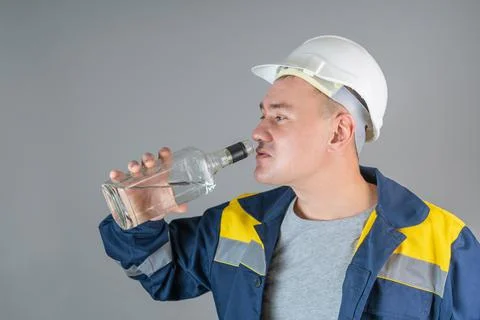 Aggressive worker in a hard hat drinks strong alcohol from a glass bottle, cl Stock Photos