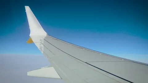 Aircraft wing view through airplane window over soft cloud layer. Bright blue Stock Footage 342334443