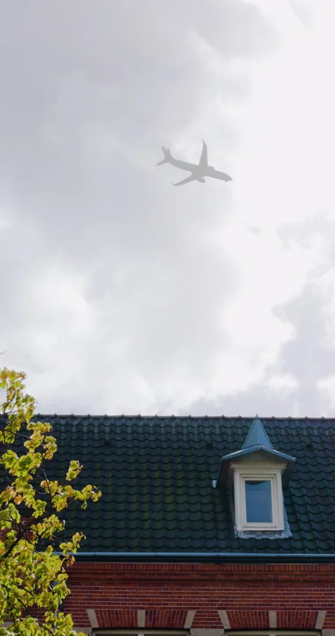 An airplane gracefully crosses a cloudy gray sky above a traditional tiled roof Vídeo Stock 322623136