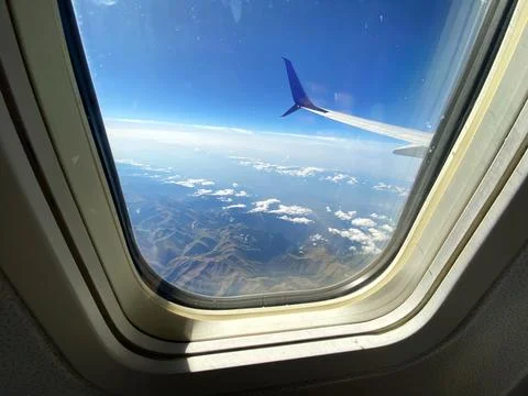 Airplane window view showcasing wing, clouds, and mountainous landscape under a Stock Photos