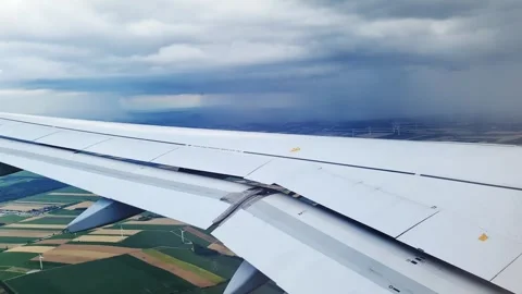 Airplane wing with deployed flaps soaring over cultivated land and wind turbines Stock Footage 347030392
