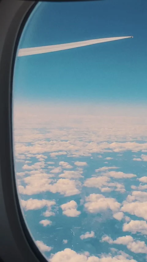 Airplane wing visible through window above fluffy clouds and blue sky, Vidéo 329201818