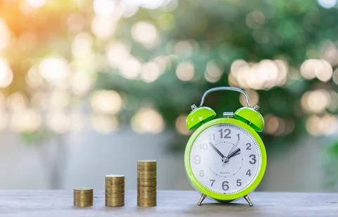 Alarm clock and multiple stacks of coins on out of focus green tree backgroun Stock-Fotos