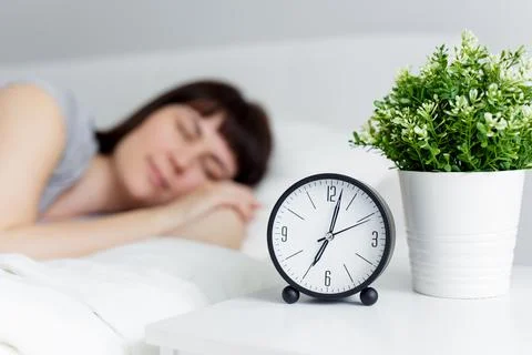 Alarm clock on bedside table and beautiful woman sleeping in bed at home Stock Photos